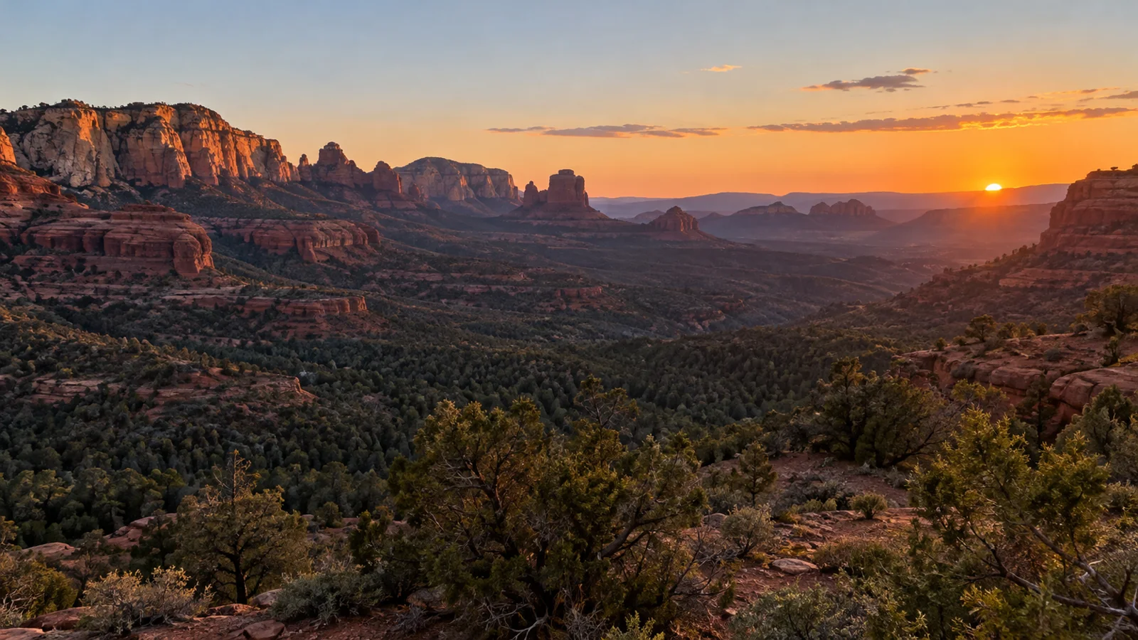 Evening sun near the horizon beyond layered red-rock mesas and juniper-covered canyon country